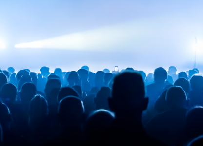 Crowd of people silhouetted in blue lighting at a concert or live event with bright stage lights in the background