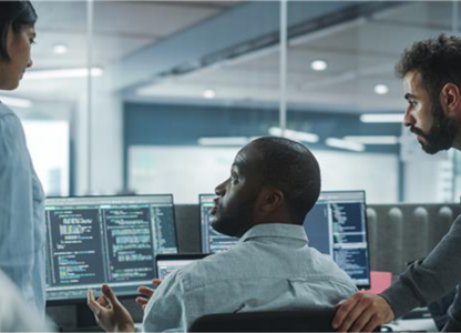 Team of IT professionals collaborating at multiple computer screens displaying code in a modern office