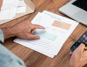 Person reviewing bills and using a calculator app on a smartphone while sitting at a table with documents and a laptop