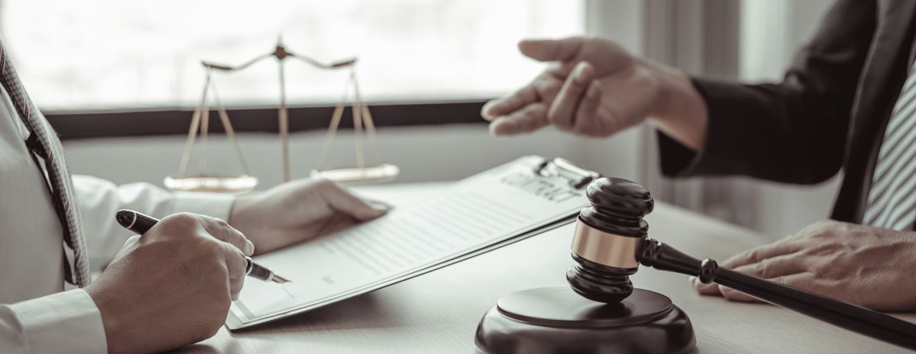 Lawyer reviewing and signing documents on a clipboard as another person gestures during a legal consultation
