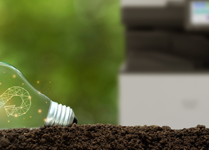 Light bulb lying on soil with a circular sustainability symbol glowing inside, with a blurred office printer in the background