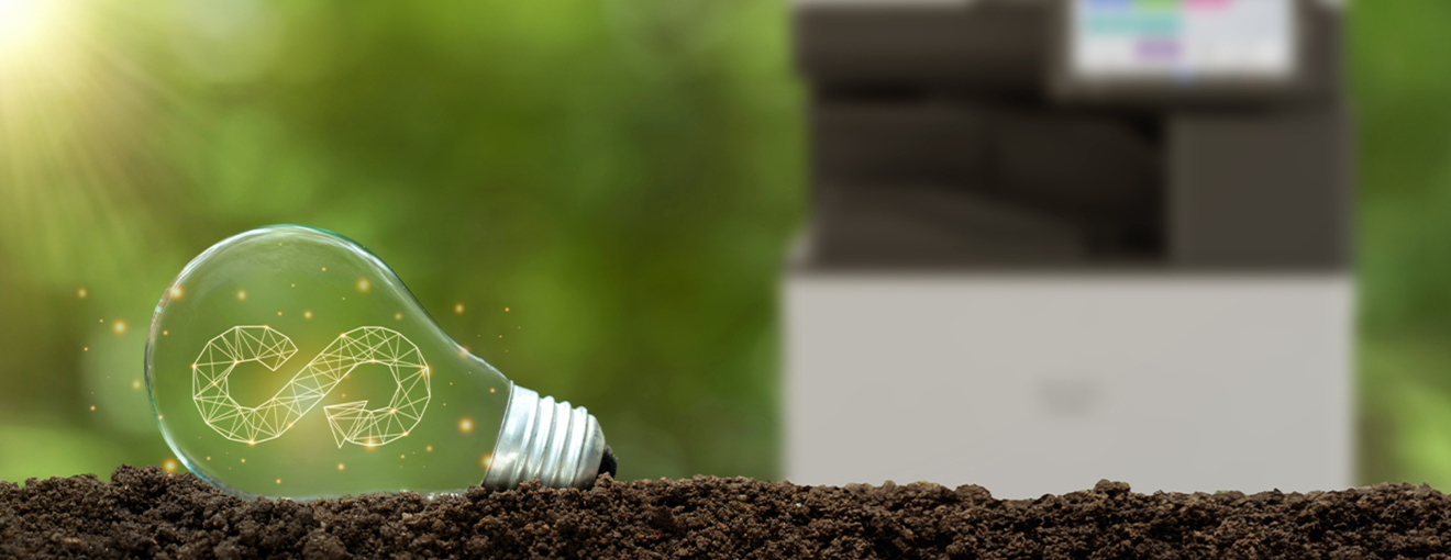Light bulb lying on soil with a circular sustainability symbol glowing inside, with a blurred office printer in the background